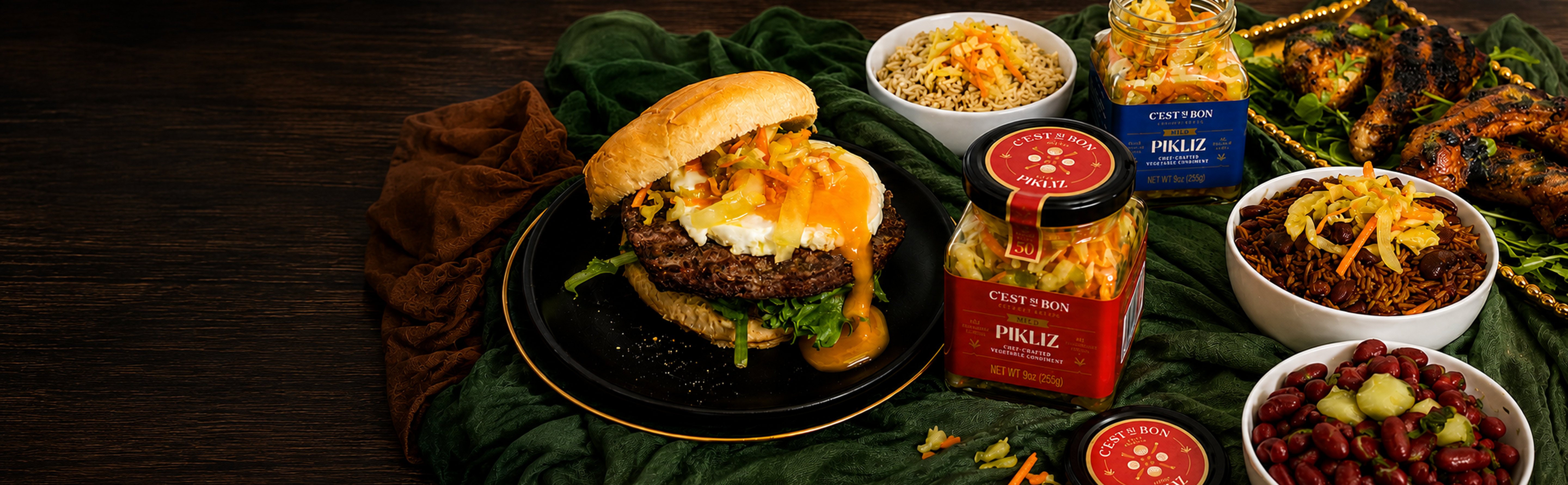 A gourmet burger with melted cheese and fresh lettuce on a black plate, styled with various side dishes and two jars of Caviar & Co. Vanilla Spice Sauce on a dark green cloth over a dark wooden table.