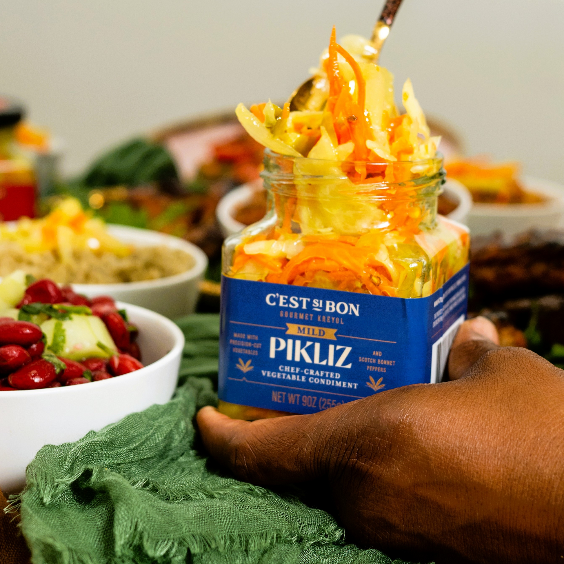 Jar of Pikliz vegetable condiment held by a hand with a blurred background of food.
