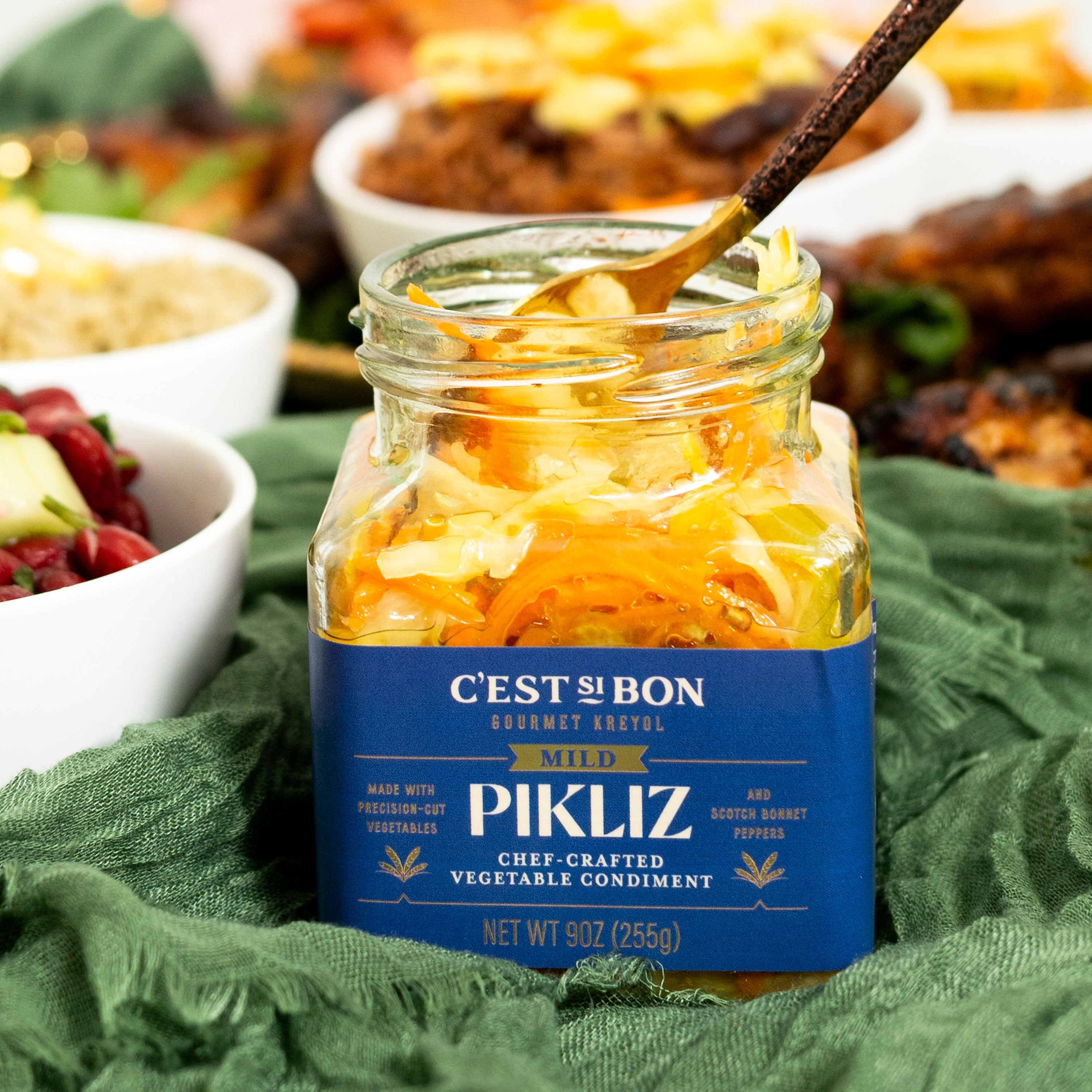 Jar of C'est Bon Pikliz vegetable condiment on a green cloth with blurred food items in the background