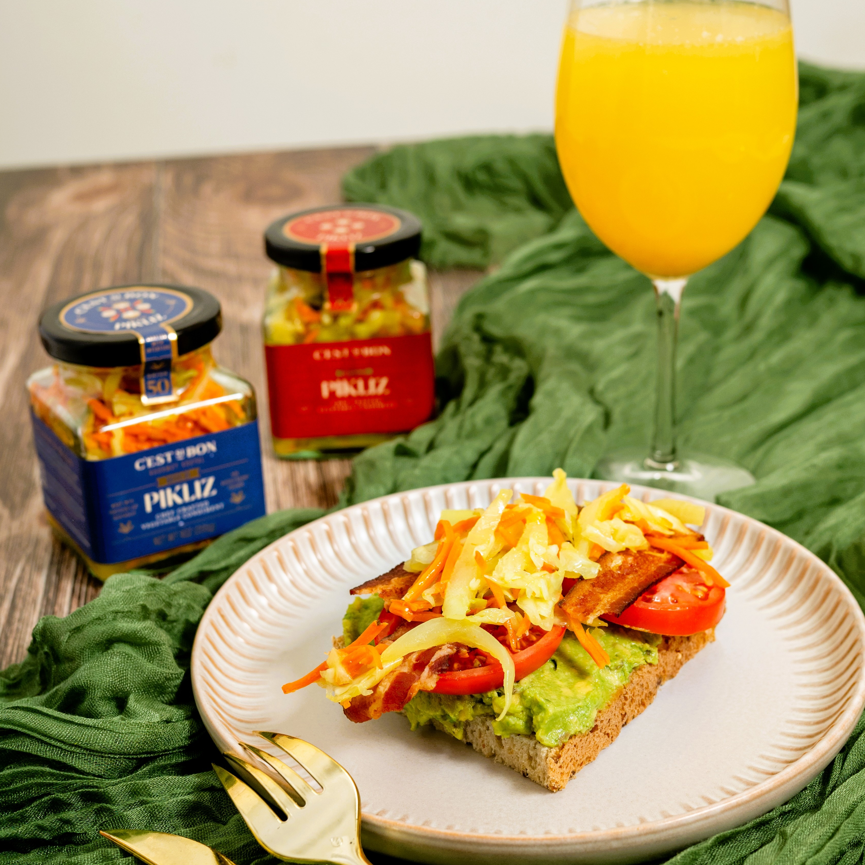 Plate of avocado toast with toppings, glass of orange juice, and jars of Pikliz on a wooden surface.