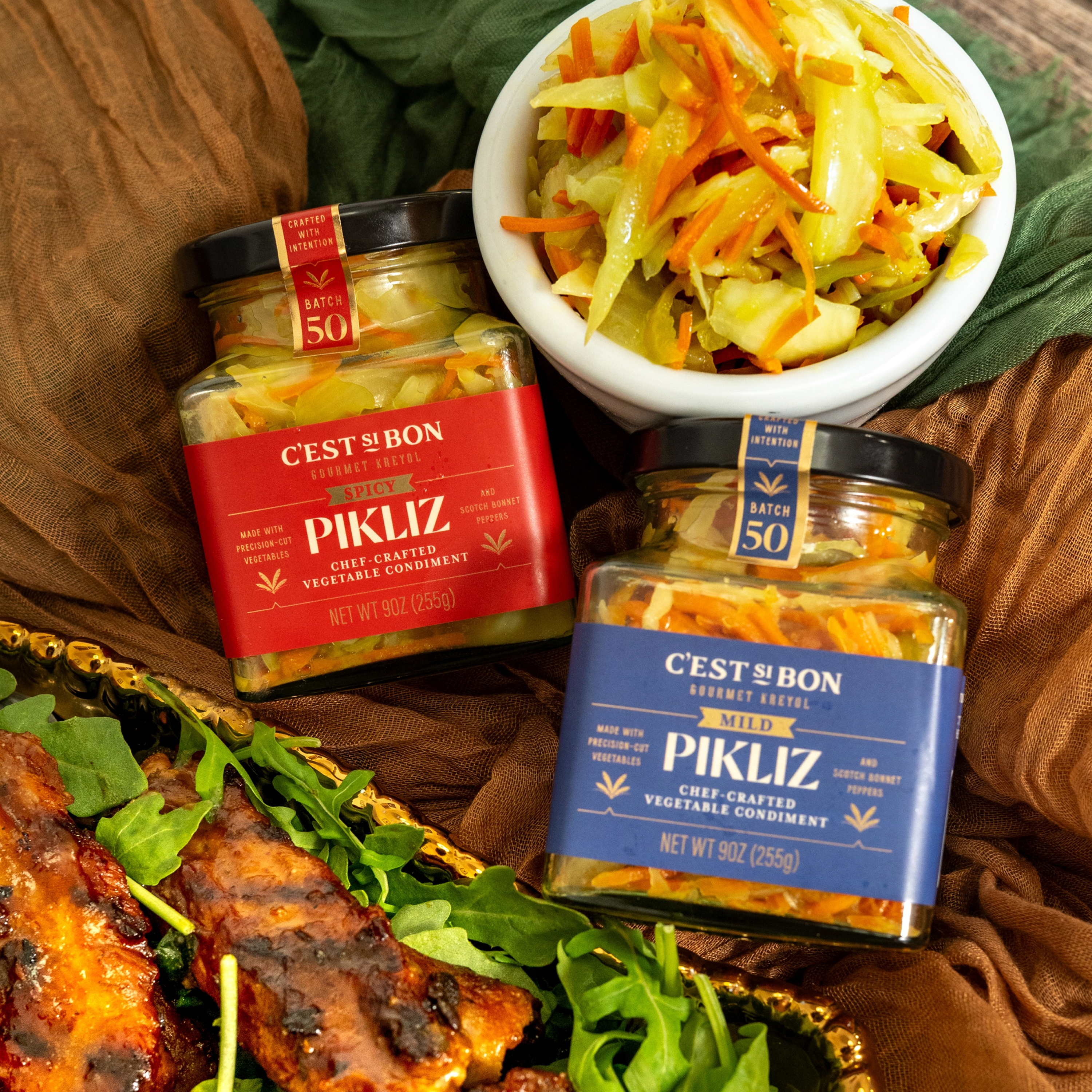 Two jars of 'C'est du Bon' Pikliz vegetable condiment with a bowl of salad and grilled food on a rustic background.
