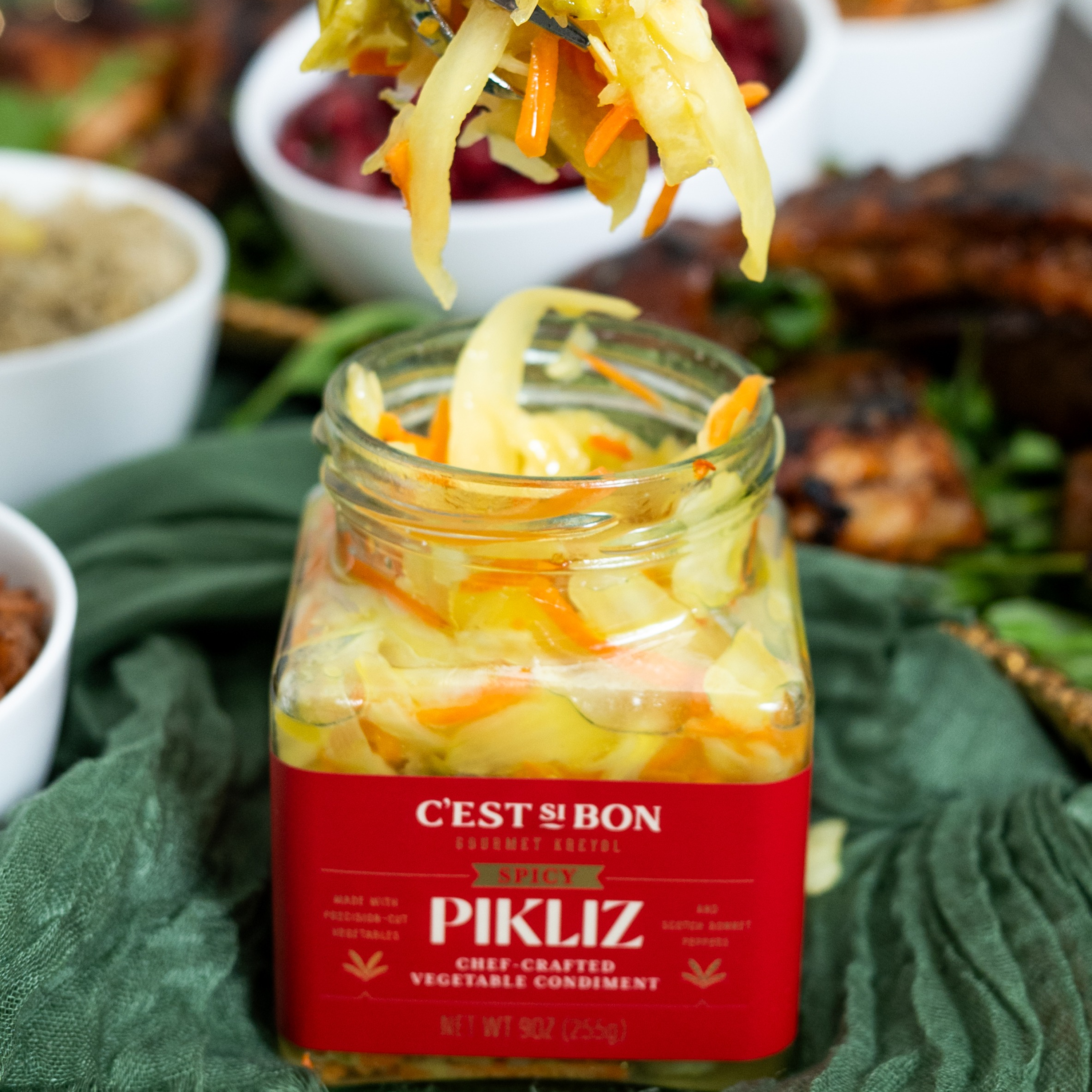 Jar of Pikliz vegetable condiment with a red label on a green surface, surrounded by other food items.